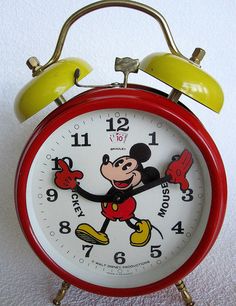 a red alarm clock with mickey mouse on it's face and yellow bell stands in front of a white wall