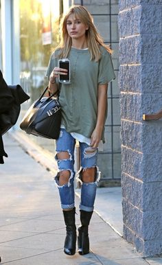 a woman with ripped jeans and black boots is holding a cell phone while walking down the street