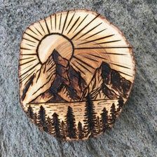 a wooden brooch with mountains and trees in the background, on a gray surface