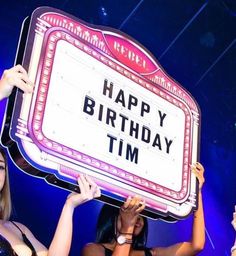 two women hold up a sign that says happy birthday tim in front of their faces