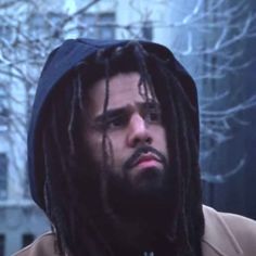 a man with dreadlocks and a hoodie looks off into the distance while standing in front of a building