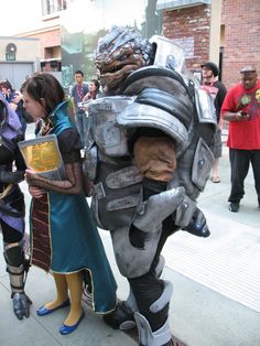 two people dressed in costumes standing next to each other on the sidewalk while others look on