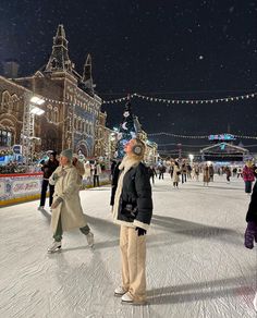 people skating on an ice rink at night with christmas lights strung across the street and buildings in the background