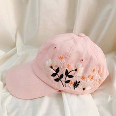 a pink baseball cap with embroidered flowers on it sitting on a white cloth covered bed