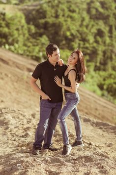 a man and woman standing on top of a dirt hill next to each other with trees in the background