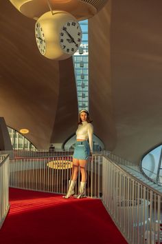 a woman is standing on a red carpet in front of a large clock hanging from the ceiling