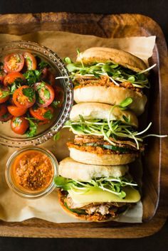 a wooden tray topped with sandwiches covered in lettuce and tomato slices next to a bowl of salsa