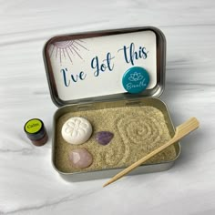 an open tin with sand and rocks in it next to two wooden chopsticks