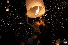 a man and woman kissing under a paper lantern in the sky with lights all around them