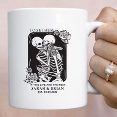 a person holding a white coffee mug with a skeleton on it's side and an engagement ring in their left hand