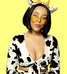 a woman wearing glasses and a cow print top with horns on her head, standing in front of a yellow background