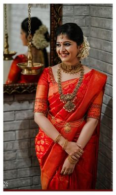 a woman in an orange and red sari standing next to a brick wall with a mirror