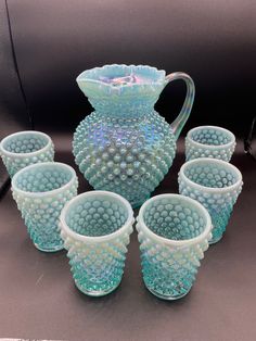 a set of six glass cups and a pitcher on a black tablecloth with other items in the background