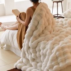 a woman sitting on top of a bed covered in a fluffy blanket and holding a book