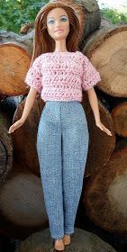 a doll is standing in front of logs wearing blue pants and a pink top with crocheted details