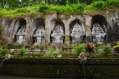 an old stone structure with statues and plants growing on the side of it, surrounded by greenery