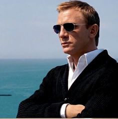 a man wearing sunglasses standing in front of the ocean with his arms crossed and looking off into the distance