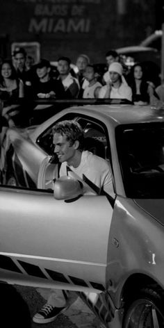 a black and white photo of a man driving a car with people in the background