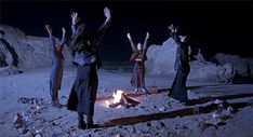 four people standing around a campfire with their hands in the air and arms raised