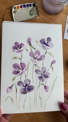 a person holding up a card with watercolors on it and purple flowers in the background