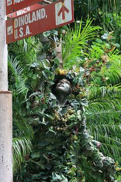 there is a statue made out of plants and street signs on the pole in front of trees