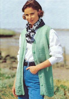 a woman standing on the beach wearing a green vest and blue jeans with her hands in her pockets