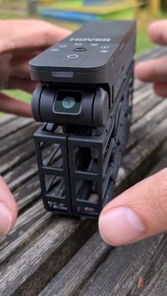 someone is holding a small camera in their hands on a wooden table outside with grass and trees behind them