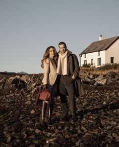 a man and woman standing on rocks in front of a white house with a red handbag