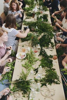 a group of people sitting around a long table covered in green plants and plates filled with food