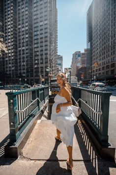 a woman in a white dress is walking across a bridge with tall buildings behind her