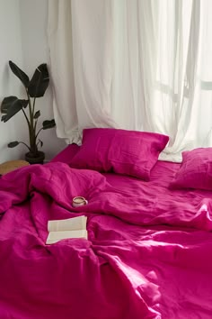 an unmade bed with pink sheets and pillows in front of a white curtained window