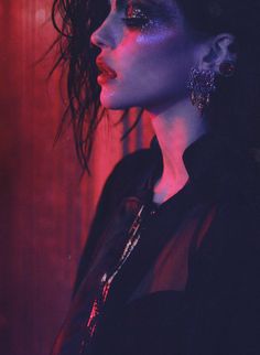 a woman with dark hair and blue makeup is wearing silver earrings in front of a red background