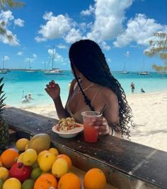 a woman sitting at a table with fruit and drinks in front of her on the beach