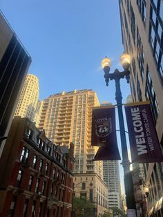 a street light in the middle of a city with tall buildings on either side and a welcome sign hanging from it's pole