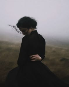 a woman in a black dress standing on a foggy field with her hair blowing in the wind