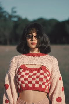 a woman with black hair wearing a red and white knitted crop top, standing in a field