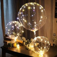 three clear balloons with lights on them sitting on a table next to flowers and books