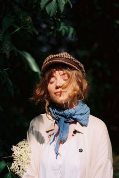 a woman wearing a hat and scarf standing in front of some trees with her eyes closed