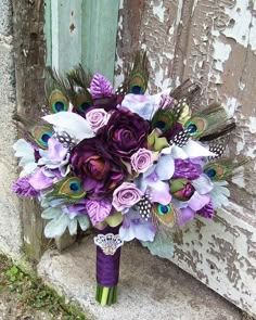 a bridal bouquet with purple flowers and peacock feathers is sitting on the ground next to an old door