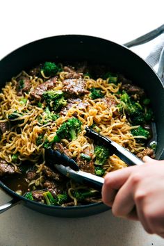 someone is holding tongs over some noodles and broccoli in a skillet