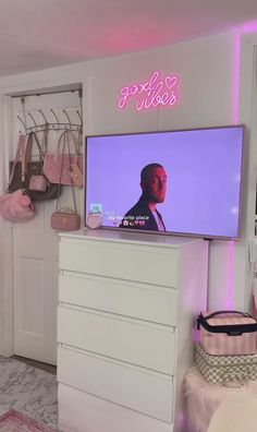 a flat screen tv mounted to the side of a white dresser next to a pink neon sign