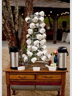 a wooden table topped with lots of white cups and saucers next to a tree