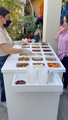 two women standing at a table with food on it and one woman wearing a face mask