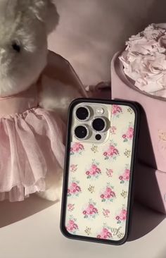 a white teddy bear wearing a pink dress next to a cell phone case with flowers on it