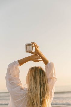 a woman standing on top of a beach holding up a cup in front of her head