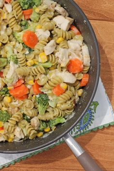 a skillet filled with pasta and vegetables on top of a wooden table next to a napkin