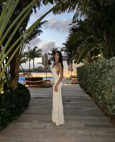 a woman in a white dress standing on a wooden walkway next to palm trees and water