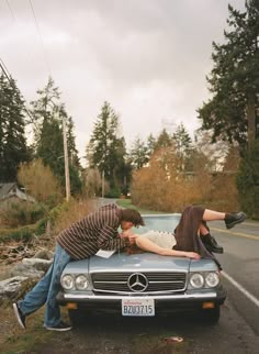 two people leaning on the hood of a car in front of a tree lined road