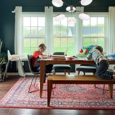 two people sitting at a table in a living room with blue walls and white windows