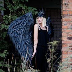 a woman with long blonde hair standing next to a black angel wings sculpture in front of a brick wall